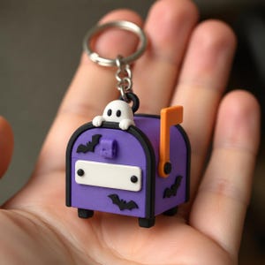May include: A purple mailbox keychain featuring a small white ghost perched on top. The mailbox has a white rectangular front panel, a bright orange flag on the side, and black bat silhouettes decorating the purple exterior surfaces.
