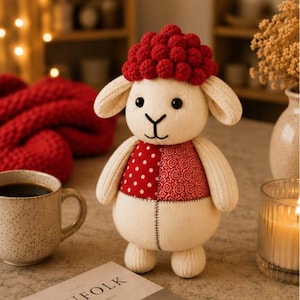 May include: A plush sheep toy with a cream-colored body, floppy ears, and black embroidered facial features. The sheep wears a textured red crocheted cap and a red vest featuring a split design of white polka dots and a red swirling pattern.