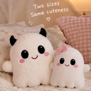 Può includere: Due peluche bianchi a forma di fantasma con occhi neri, guance rosa e bocche sorridenti. Il più grande ha corna nere, il più piccolo un fiocco rosa. Il testo sopra recita: Two sizes Same cuteness con un piccolo simbolo a forma di cuore.