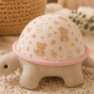 May include: A stuffed turtle toy with a beige fabric body and a dome-shaped shell featuring a cream background with a repeating pattern of brown teddy bears, white rabbits, and small pink and yellow flowers, finished with a soft pink felt trim.