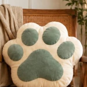 May include: A plush, cream-coloured throw cushion shaped like an animal paw. The cushion features four rounded toe pads and one large, central heart-shaped pad, all rendered in a soft, muted sage green colour with a textured, velvety fabric finish.