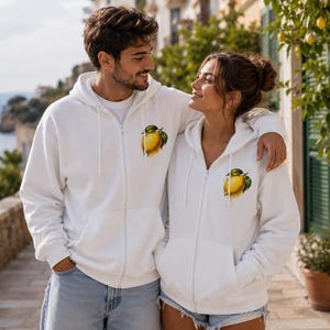 Op de afbeelding: Een witte hoodie met rits en een kleine, gedetailleerde afbeelding van een enkele gele citroen met groene bladeren op de linkerborst. Het kledingstuk is voorzien van een capuchon met trekkoord, een ritssluiting en twee voorzakken.