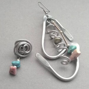 May include: A pair of silver earrings with a unique design. One earring is a simple spiral, while the other is a larger, more elaborate design with multiple loops and a small gold bead. Both earrings feature turquoise and pink stones.
