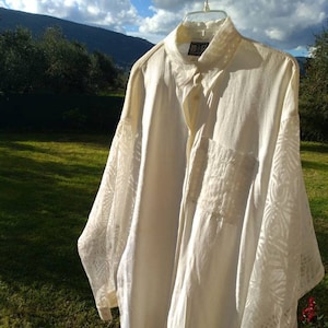 May include: A white long-sleeved button-down shirt with a lace overlay on the sleeves and a small pocket on the left chest. The shirt is hanging on a clothesline outdoors.