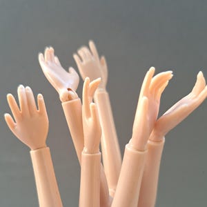 May include: Several light-coloured plastic doll arms and hands, with visible joints. The hands are in different positions, some open and some partially closed, set against a plain background.