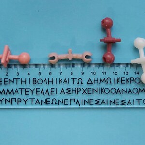 May include: Four plastic toy pieces, two pink, one white, and one red, with round ends and a connecting piece in the middle. The pieces are arranged on a blue surface with a ruler marked in centimeters.
