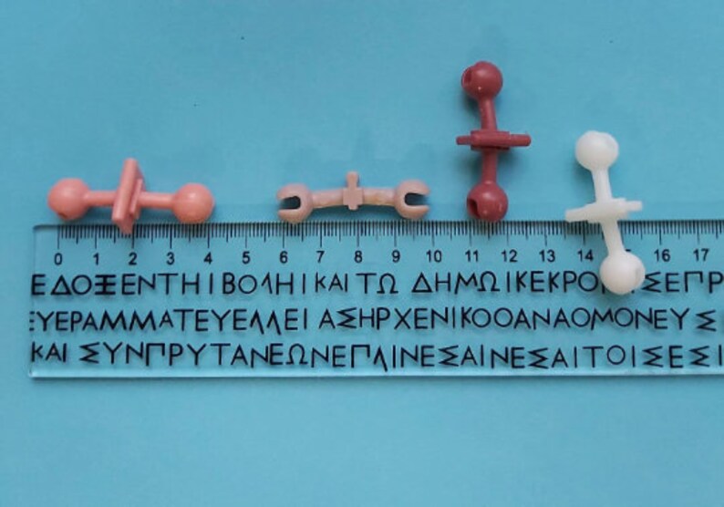 May include: Four plastic toy pieces, two pink, one white, and one red, with round ends and a connecting piece in the middle. The pieces are arranged on a blue surface with a ruler marked in centimeters.