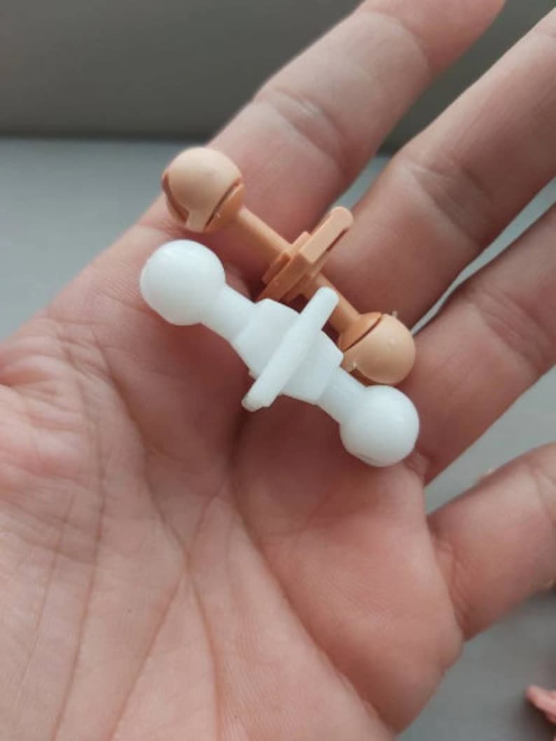 May include: Two doll joint connectors, one white and one beige, with ball-shaped ends. The connectors are designed for attaching doll limbs to the body.