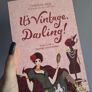 Può includere: Copertina di un libro con il titolo "It's Vintage, Darling!" in corsivo bordeaux. L'illustrazione presenta una donna in abiti vintage, con uno specchio e un rossetto, insieme a un manichino e un servizio da tè. Lo sfondo è un motivo floreale rosa.