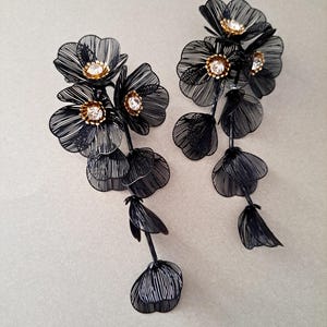 Puede incluir: Un par de pendientes florales de alambre negro. Cada pendiente presenta múltiples racimos de flores con centros de cristal transparente y detalles dorados. Los pendientes tienen un diseño colgante con delicados pétalos en capas.