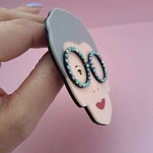 May include: A gray and white resin brooch of a woman wearing round glasses with iridescent rhinestones. The brooch has a black outline.