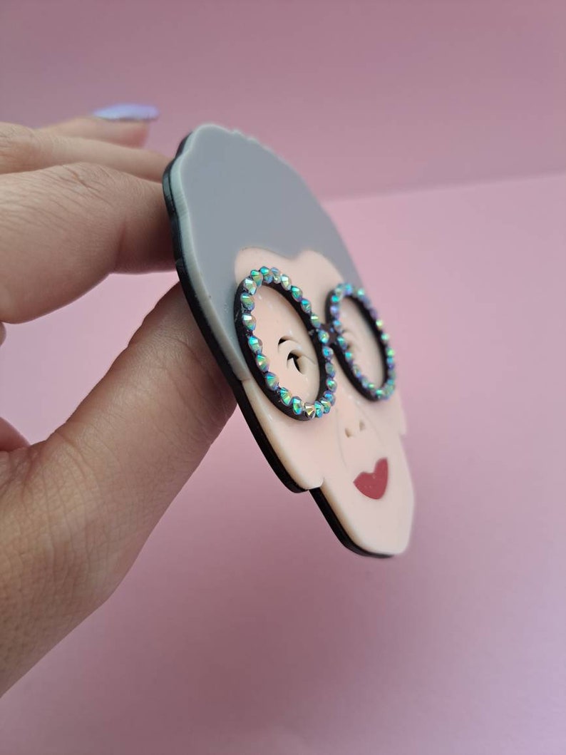 May include: A gray and white resin brooch of a woman wearing round glasses with iridescent rhinestones. The brooch has a black outline.