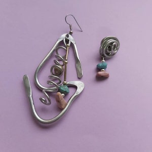 May include: A pair of silver earrings with a unique design. One earring features a large, abstract shape with a spiral, beads, and a small turquoise stone. The other earring is a smaller, round shape with a turquoise stone.