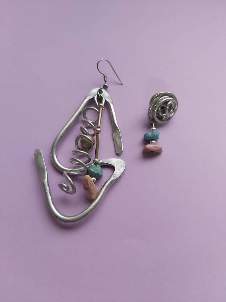 May include: A pair of silver earrings with a unique design. One earring features a large, abstract shape with a spiral, beads, and a small turquoise stone. The other earring is a smaller, round shape with a turquoise stone.