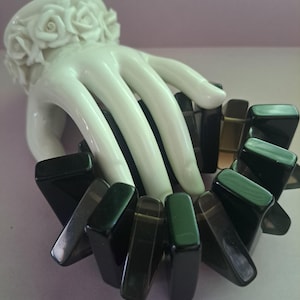 Può includere: Un bracciale elastico composto da segmenti rettangolari in pietra nera lucida e grigio scuro traslucido. Il bracciale è esposto su una scultura a mano in ceramica bianca con un polsino decorato con rose bianche scolpite.