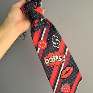 May include: A novelty necktie with a navy blue polka dot and red striped pattern. The tie features patches of a smiley face, dollar sign, red lips, strawberry, and the word "oops!" in a speech bubble. A silver chain is attached.