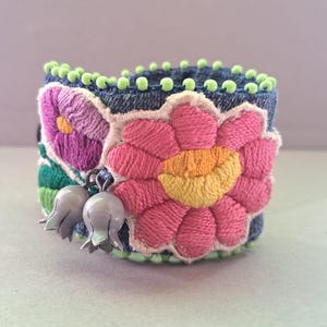 Puede incluir: Brazalete bordado con base de mezclilla, con una gran flor rosa con centro amarillo y una flor morada más pequeña. La pulsera está adornada con cuentas verdes y dijes de campana plateados, mostrando un diseño vibrante y artesanal.