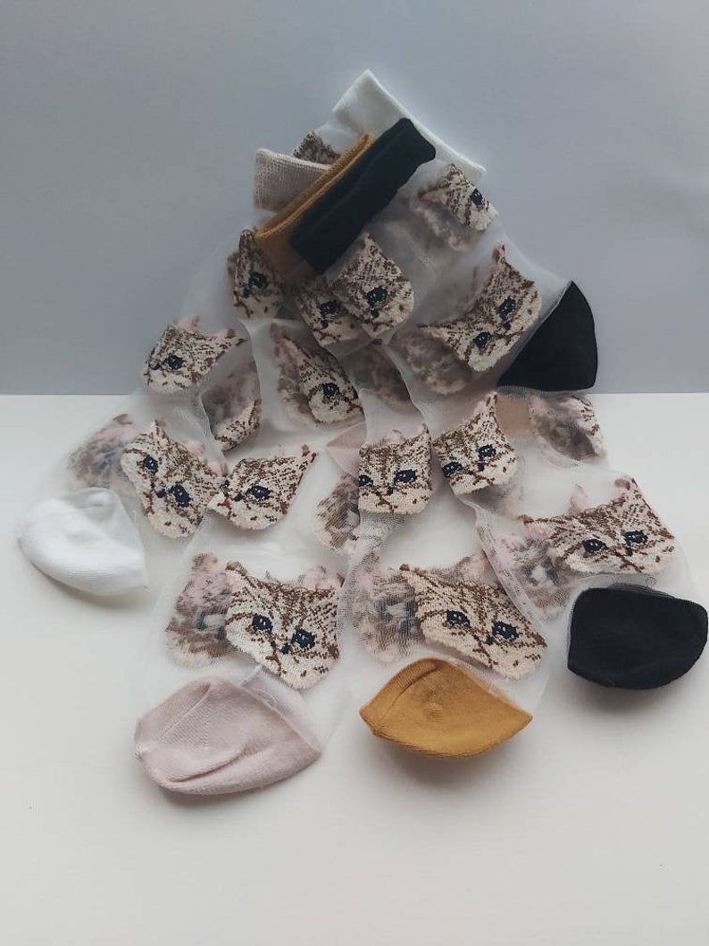 May include: A set of sheer socks with a cat face design. The socks are white with a brown cat face printed on them. The socks are available in a variety of colors, including white, pink, and yellow.