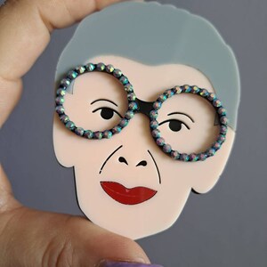 May include: A light pink acrylic brooch with a cartoon face, gray hair, and black framed glasses with iridescent rhinestones. The face has red lips and a small nose.