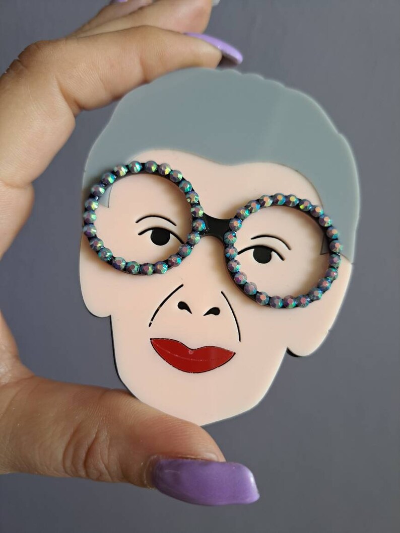 May include: A light pink acrylic brooch with a cartoon face, gray hair, and black framed glasses with iridescent rhinestones. The face has red lips and a small nose.
