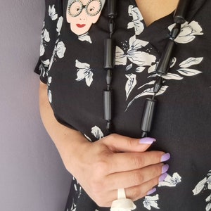May include: A black blouse with a white floral pattern. The blouse features a large, white, ceramic brooch of a woman wearing glasses. The woman is wearing a black necklace with large, black, cylindrical beads.