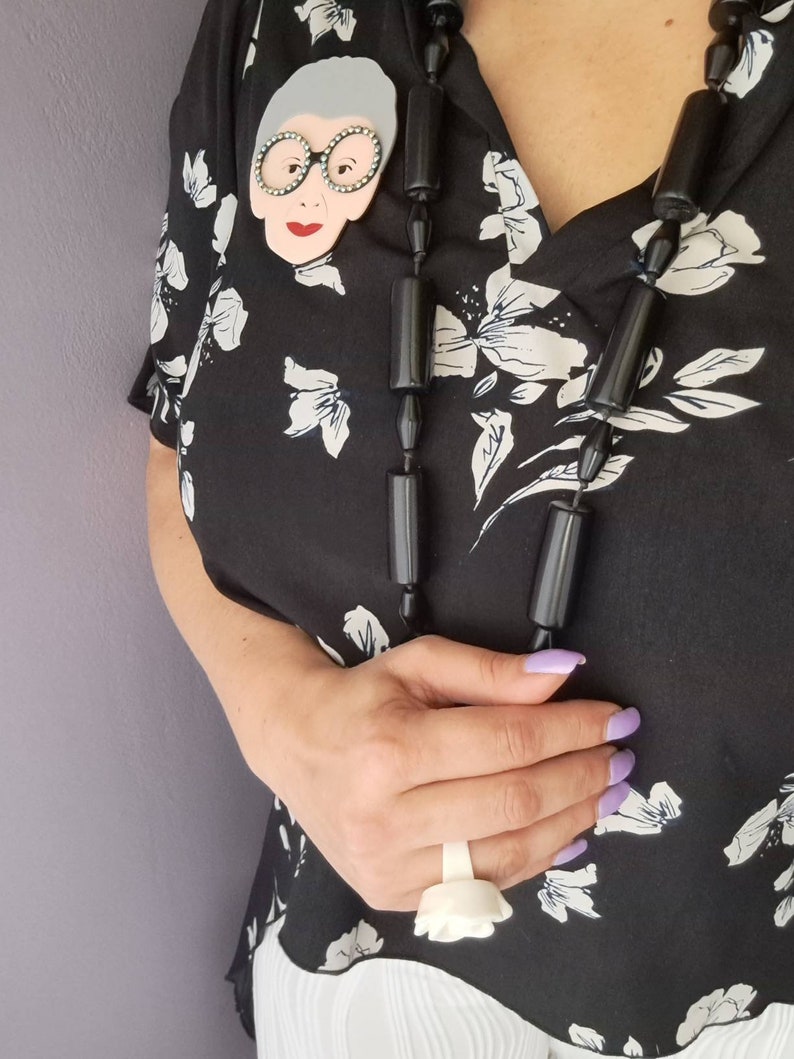 May include: A black blouse with a white floral pattern. The blouse features a large, white, ceramic brooch of a woman wearing glasses. The woman is wearing a black necklace with large, black, cylindrical beads.