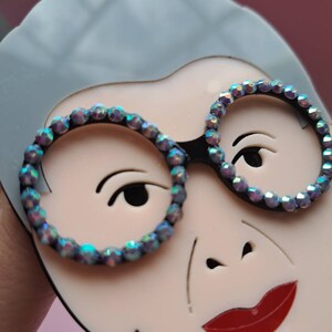 May include: A close-up of a white face with gray hair, wearing round glasses with iridescent rhinestones. The face has a red mouth and a black nose.