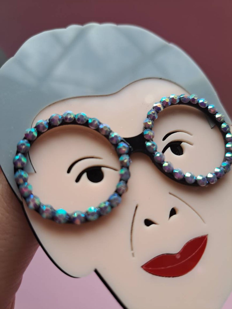 May include: A close-up of a white face with gray hair, wearing round glasses with iridescent rhinestones. The face has a red mouth and a black nose.