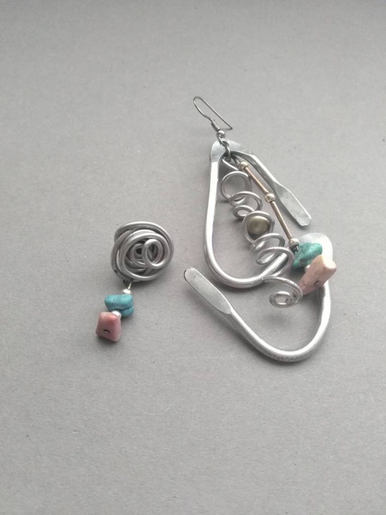 May include: A pair of silver earrings with a unique design. One earring is a simple spiral, while the other is a larger, more elaborate design with multiple loops and a small gold bead. Both earrings feature turquoise and pink stones.