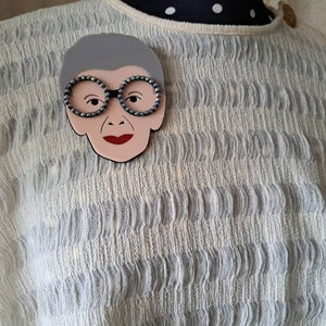 May include: A gray and white brooch featuring a cartoon face with black glasses and red lips. The brooch is made of plastic and has a shiny finish.