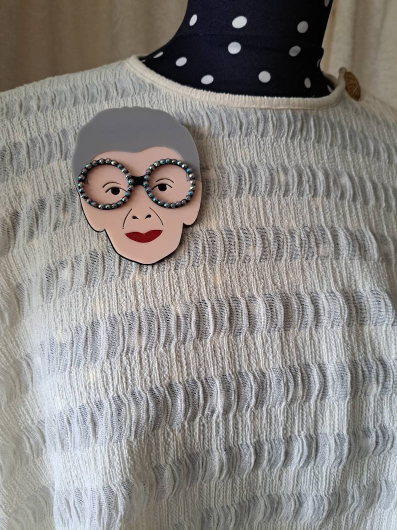 May include: A gray and white brooch featuring a cartoon face with black glasses and red lips. The brooch is made of plastic and has a shiny finish.