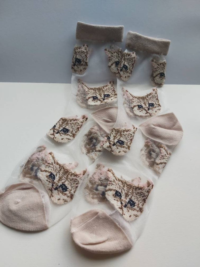 May include: A pair of sheer socks with a cat face pattern. The socks are light pink and have a brown cat face design.