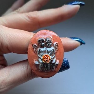 May include: A peach-coloured oval ring with a cameo of two angels holding a flower. The angels are sculpted in a grey colour and the flower is orange.