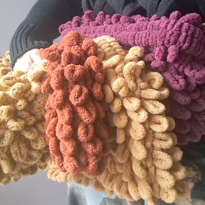 Puede incluir: Una colección de mantas hechas a mano y texturizadas en varios colores, incluyendo óxido, beige y morado. Las mantas tienen un diseño único con bucles, creando una apariencia suave y afelpada. La imagen muestra la calidad táctil y las variaciones de color de las mantas.