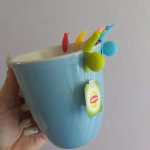 Puede incluir: Una taza de cerámica azul con una bolsita de té colgando del borde. La bolsita de té es una bolsita de té de la marca Lipton. Hay cinco infusores de té de caracol de silicona de colores unidos a la taza.