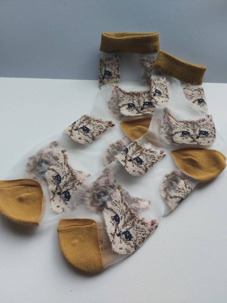 May include: A pair of sheer white socks with a mustard yellow cuff. The socks feature a repeating pattern of brown and white cat faces.