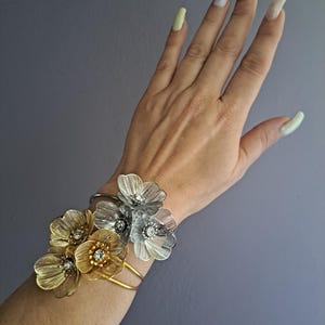 May include: A gold and silver floral bracelet with rhinestones. The bracelet is made of metal and features three flowers, two gold and one silver.