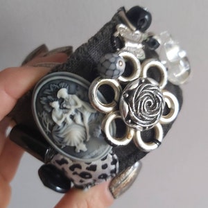 Può includere: Un braccialetto in tessuto nero con decorazioni argentate e nere, tra cui un cammeo, un fiore con una rosa al centro e una toppa di tessuto con stampa leopardata.
