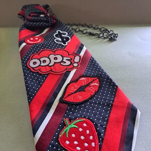 May include: A novelty necktie with a red, white, and navy blue striped pattern, adorned with embroidered patches. Patches include a strawberry, lips, a dollar sign, a smiley face, and the word "oops!" in a speech bubble.