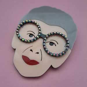 May include: A white and gray resin brooch depicting a woman wearing glasses with iridescent rhinestones. The woman has a red mouth and gray hair.