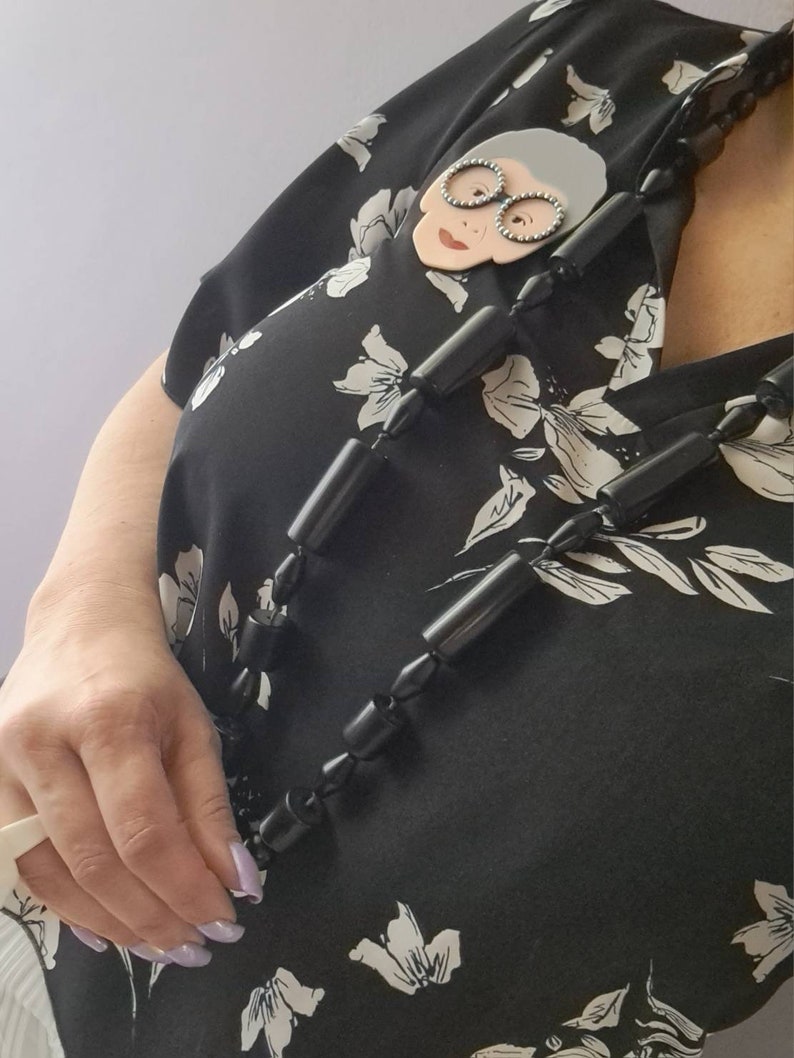 May include: A black and white floral blouse with a black beaded necklace. The necklace features a large, white, cartoon-style face with glasses.