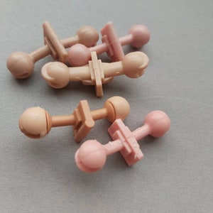 May include: A group of six plastic doll head connectors in two colors: light brown and light pink. The connectors have a small ball on one end and a flat, rectangular piece on the other end.