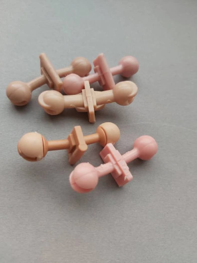 May include: A group of six plastic doll head connectors in two colors: light brown and light pink. The connectors have a small ball on one end and a flat, rectangular piece on the other end.