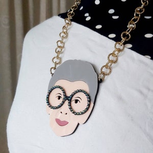 May include: A gold chain necklace with a gray and pink portrait pendant featuring a woman wearing round glasses with iridescent rhinestones.