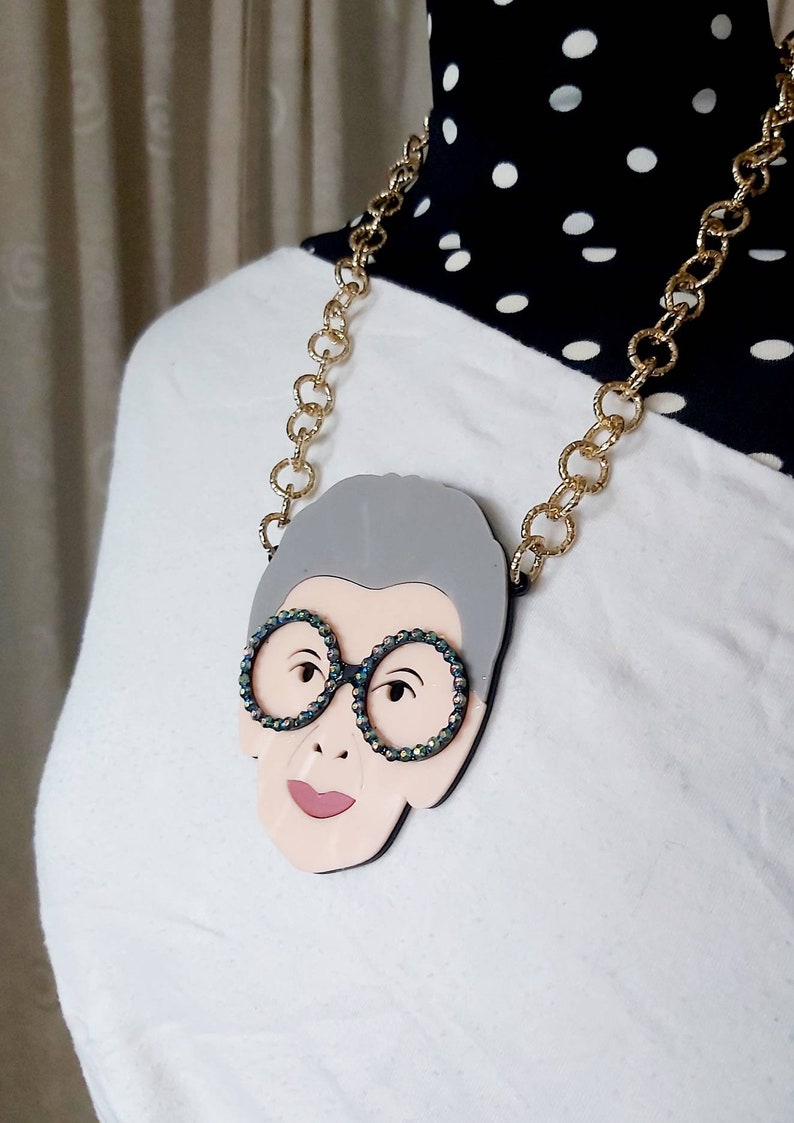 May include: A gold chain necklace with a gray and pink portrait pendant featuring a woman wearing round glasses with iridescent rhinestones.