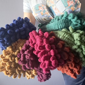 Puede incluir: Una colección de artículos de hilo de bucle hechos a mano en varios tonos, incluyendo azul, dorado, rosa, burdeos, verde y naranja. El hilo suave y texturizado está dispuesto en forma de abanico, mostrando la gama de colores y la calidad táctil del material.