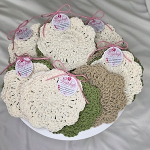 May include: A collection of round, scalloped-edge crochet coasters in cream, sage green, and tan. Each coaster features a circular paper tag with the text Majicleigh Crochet Co. attached by a thin pink string tied in a small bow at the top.