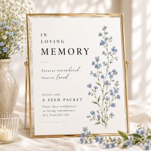 May include: A gold-framed sign with white background featuring black text that reads IN LOVING MEMORY, Forever remembered, forever loved, and PLEASE TAKE A SEED PACKET, Plant these wildflowers in loving remembrance of a beautiful life, with blue flowers.