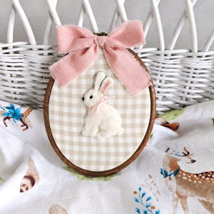 May include: An oval embroidery hoop frame containing a beige and white gingham fabric background. A small, white, three-dimensional felt rabbit with a tiny pink ribbon around its neck is centered. A frayed, dusty pink fabric bow sits at the top.