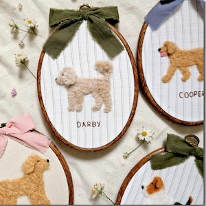 May include: Oval embroidery hoops with dog portraits. Each hoop features a fabric background with a vertical stripe pattern, a brown wooden frame, and a decorative bow at the top. The dogs are made of textured yarn and the names "Darby" and "Cooper" are embroidered.